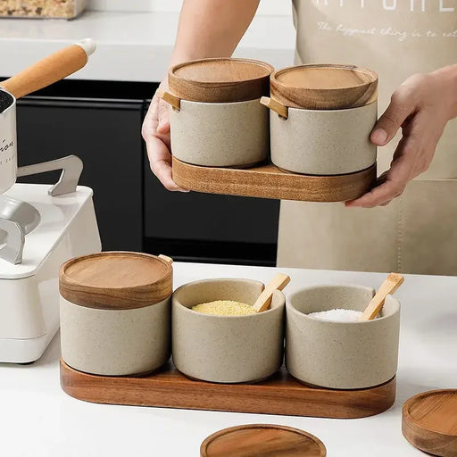 Person holding a set of ceramic containers with wooden lids on a kitchen counter.